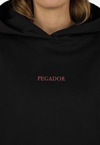 Čierna mikina s kapucňou s jemnou textúrou, so stredovo umiestneným, ružovým potlačeným textom "PEGADOR" na hrudníku. Kapucňa je navrhnutá s jednoduchým obrysom.