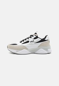 Zapatilla deportiva blanca y beige con paneles de malla, cordones negros y una suela de goma gruesa con parte trasera redondeada y el logo de Fila.