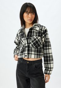 Giacca in flanella cropped in plaid bianco e nero, con chiusura frontale a bottoni, colletto e tasca. Indossata sopra un top chiaro e jeans neri.