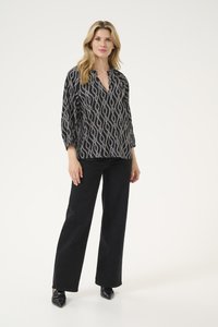 Schwarz gemusterte Bluse mit weißem geometrischen Design, V-Ausschnitt und Dreiviertelärmeln, kombiniert mit schwarzen, weit geschnittenen Jeans und High Heels.