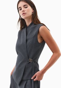 Grå ærmeløs top med indpakningsdesign, der har en sidelukning og lommer. Stoffet virker glat med en struktureret pasform.