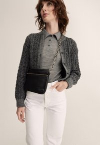 Gray knitted cardigan with textured sleeves, paired with a gray polo shirt. White high-waisted jeans and a small black leather crossbody bag.