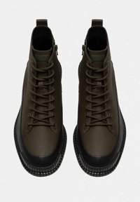 Botas tobilleras verde oliva fabricadas en cuero liso, con cordones en la parte delantera, cremalleras laterales y suela de goma negra que presenta un patrón texturizado.