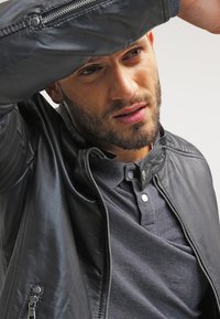 Chaqueta de cuero negra con cremallera y cierre a presión, llevada sobre una camiseta polo gris. Detalle en primer plano del cuello y la textura de la tela.