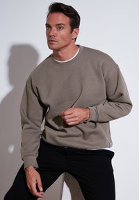 Lichtbruin sweatshirt met een losse pasvorm, ronde hals, geribbelde manchetten en een witte binnenlaag die zichtbaar is. Draagt een zwarte broek, op een lichte achtergrond.