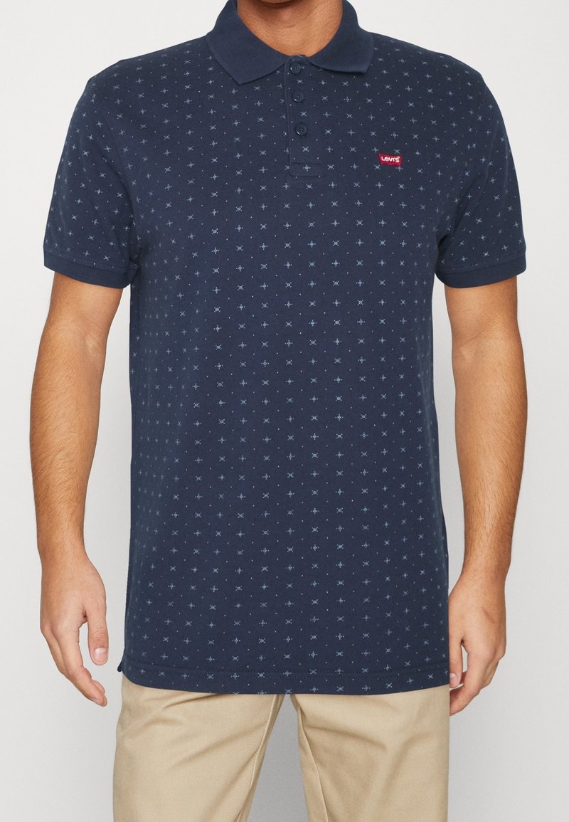 Homme portant un polo bleu marine à manches courtes avec de petits motifs en forme de croix bleu clair et un pantalon beige.