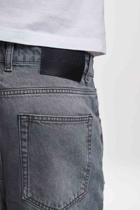 Grå denimbukser med en glatt tekstur, fem lommer og en svart skinnbelteskilt som viser merkevarenavnet i opphøyet skrift.