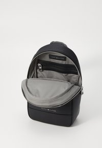 Black leather backpack with textured exterior, gray lined interior, zippered compartments, and a prominent logo label on the front.