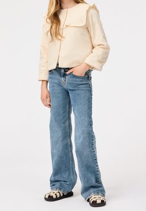 Chaqueta rizada de color beige claro con mangas largas, llevada sobre unos jeans acampanados azules. El calzado presenta un diseño tejido en negro y crema.