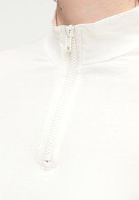 Λευκή zip-up μπλούζα φτιαγμένη από μαλακό ύφασμα, με ψηλό γιακά και φερμουάρ ενός τετάρτου μήκους με υφασμάτινη λαβή.