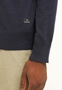 Pull en maille bleu foncé avec des poignets et un ourlet, étiquette Scotch & Soda, porté sur un pantalon beigne, montrant la main et le poignet gauche d'une personne.