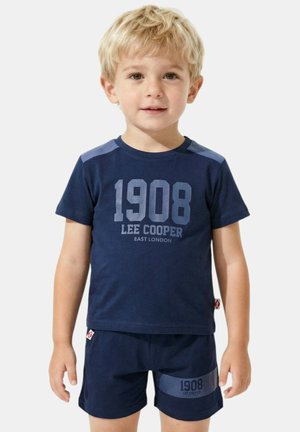 Garçon blond portant un t-shirt et un short bleu marine Lee Cooper avec le texte "1908", se tenant devant un fond clair uni.