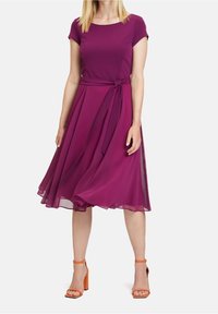 Fialové šaty s přiléhavým korzetem a krátkými rukávy, s vázacím páskem v pase. Plynulá sukně ke kolenům s průhlednou texturou. Oranžové sandály na podpatku.