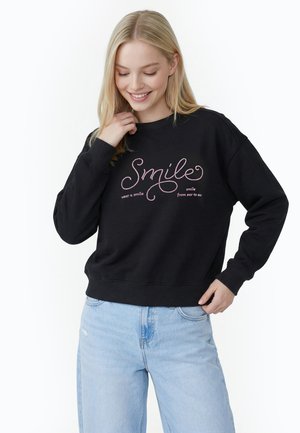 Ung kvinde smilende, iført sort sweatshirt med pink "Smile" tekst og lyseblå jeans, hånd nær krave og hofte.