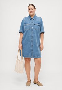 Denim shirt dress in light blue with a button-front, short sleeves, two chest pockets, and a relaxed fit, paired with a woven bag.
