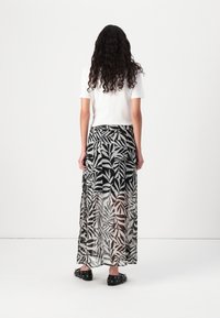 Personne aux longs cheveux bouclés portant un haut blanc à manches courtes et une jupe transparente noire et blanche à motifs de feuilles, vue de dos.