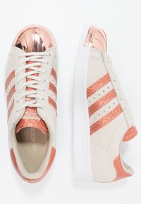 Beige sneakers with rose gold metallic shell toe and accents, featuring three rose gold stripes, white sole, and textured overlays.