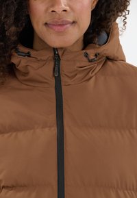 Brun oppustet jakke med lynlås, hætte og justerbare snore. Tekstureret stof med quiltet mønster og sorte hardware detaljer.