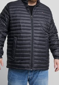 Svart vadderad jacka med horisontell söm, dragkedja framtill och sidofickor. Materialet verkar slätt med en glansig yta.
