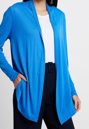 Kvinde iført en lys blå cardigan med åben front over en hvid top og sorte bukser med sort bælte, hånd i lommen.