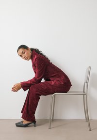 Traje de terciopelo burdeos con un blazer estructurado y pantalones de pierna ancha. Mules negros de punta. Posado sentado en una silla metálica contra una pared blanca.