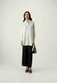 Gaiši zaļš svītrains oversize krekls ar pogu aizdari, saskaņots ar melniem izplešamiem biksēm un melniem atvērtas purngala sandalēm, turot melnu somu.