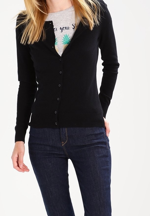 Femme portant un cardigan noir ajusté à boutons sur un t-shirt gris avec un motif et un jean bleu foncé, debout devant un fond blanc.