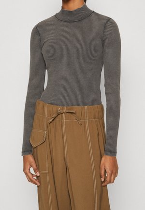 Person trägt ein eng anliegendes graues, geripptes Langarmshirt mit Rollkragen und lockere braune Hose mit Kordelzug und aufgesetzter Tasche an der Seite.