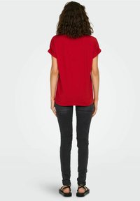 Kvinde med krøllet brunt hår iført en rød kortærmet skjorte, sorte tætsiddende jeans og sorte sandaler, stående med ryggen til på hvid baggrund.