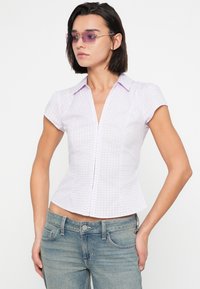 Ανοιχτό μωβ καρό πουκάμισο με εφαρμοστή γραμμή, κοντά μανίκια καπέλο και λαιμόκοψη σε σχήμα V. Λεία υφή, λεπτομέρειες στις ραφές και διακριτικό κούμπωμα με κουμπιά.