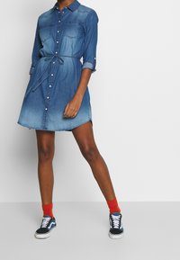 Denim-mekko, jossa on nappikiinnitys, kaksi rintataskua, kireä vyötärönauha, pitkät hihat ja liukuväri sinisissä sävyissä. Yhdistetty tummiin lenkkareihin ja punaisiin sukkiin.