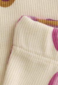 Nahaufnahme von cremefarbenem, geripptem Strickstoff mit gefaltetem Rand und abstrakten lila und braunen Mustern.