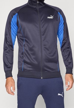 Mężczyzna noszący czarną i niebieską sportową kurtkę Puma na zamek oraz pasujące granatowe spodnie sportowe Puma, pokazany od szyi do połowy uda.