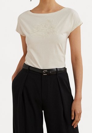 Femme portant un chemisier blanc à manches courtes avec broderie florale et un pantalon noir taille haute avec une ceinture noire, mains dans les poches.