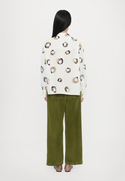 Camisa blanca con un patrón circular en colores apagados, combinada con pantalones de pana verde oliva de pierna ancha y zapatos de punta abierta multicolores.