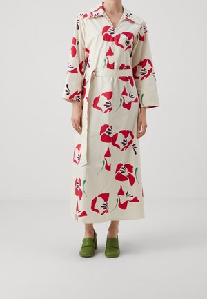 Kvinde iført en lang cremefarvet kjole med store røde blomsterprint, et bælte og grønne højhælede sko stående mod en ensartet hvid baggrund.