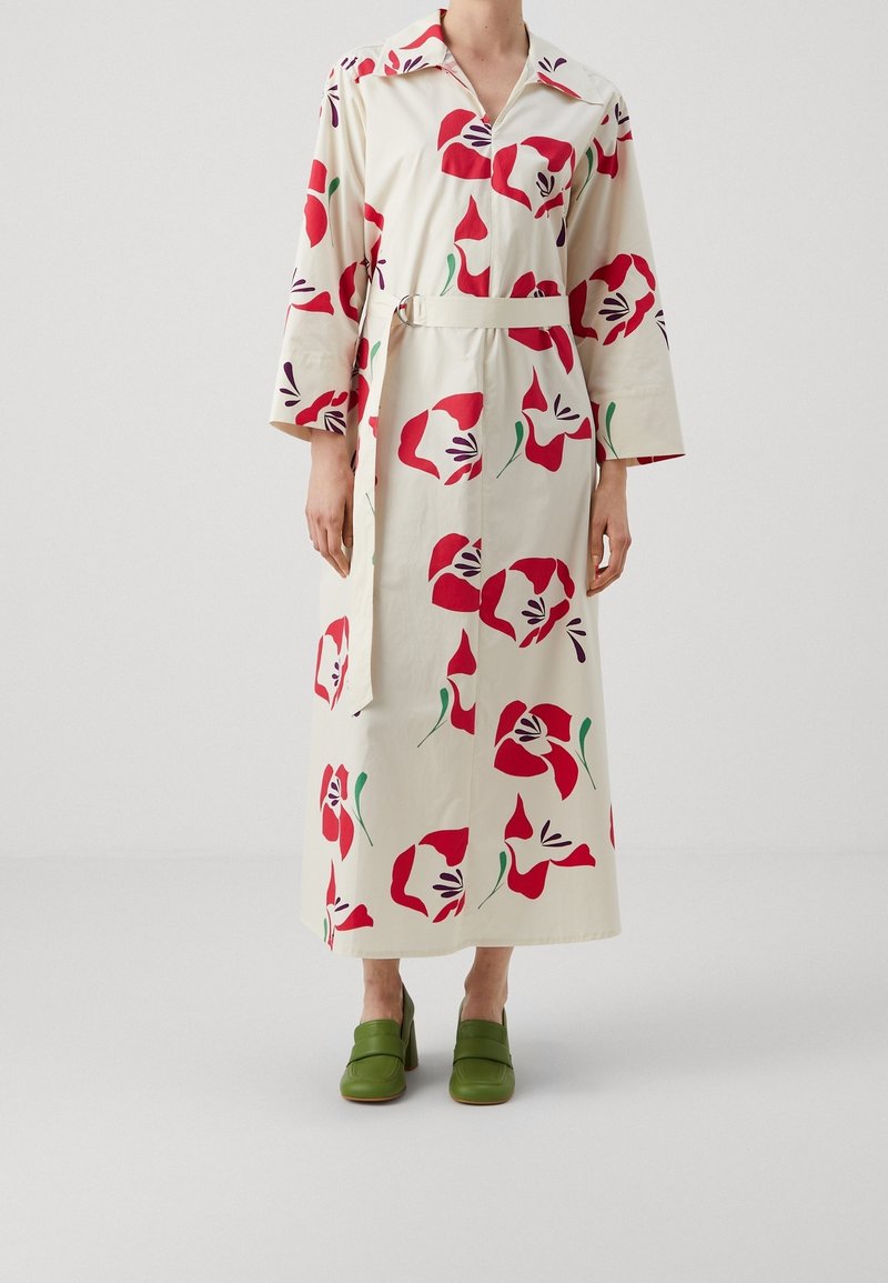 Femme portant une longue robe crème avec de grandes impressions florales rouges, une ceinture et des chaussures à talons vertes, debout devant un fond blanc uni.