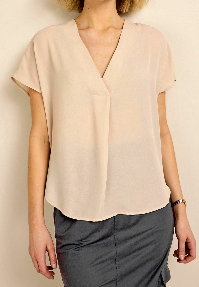 Femme portant un chemisier beige clair à manches courtes avec un décolleté en V et une jupe gris foncé, debout devant un fond beige.