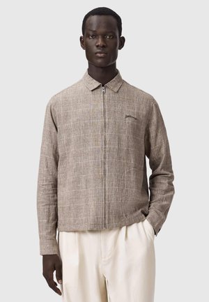 Jonge man die een beige gestructureerd shirt met rits en kraag draagt, met crèmekleurige broek, staand met één hand in de zak.