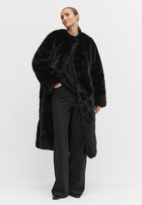 Faux fur ytterrock i svart med en lång, flödande design. Har en rundad krage och sidofickor, i kombination med figurnära svarta byxor.