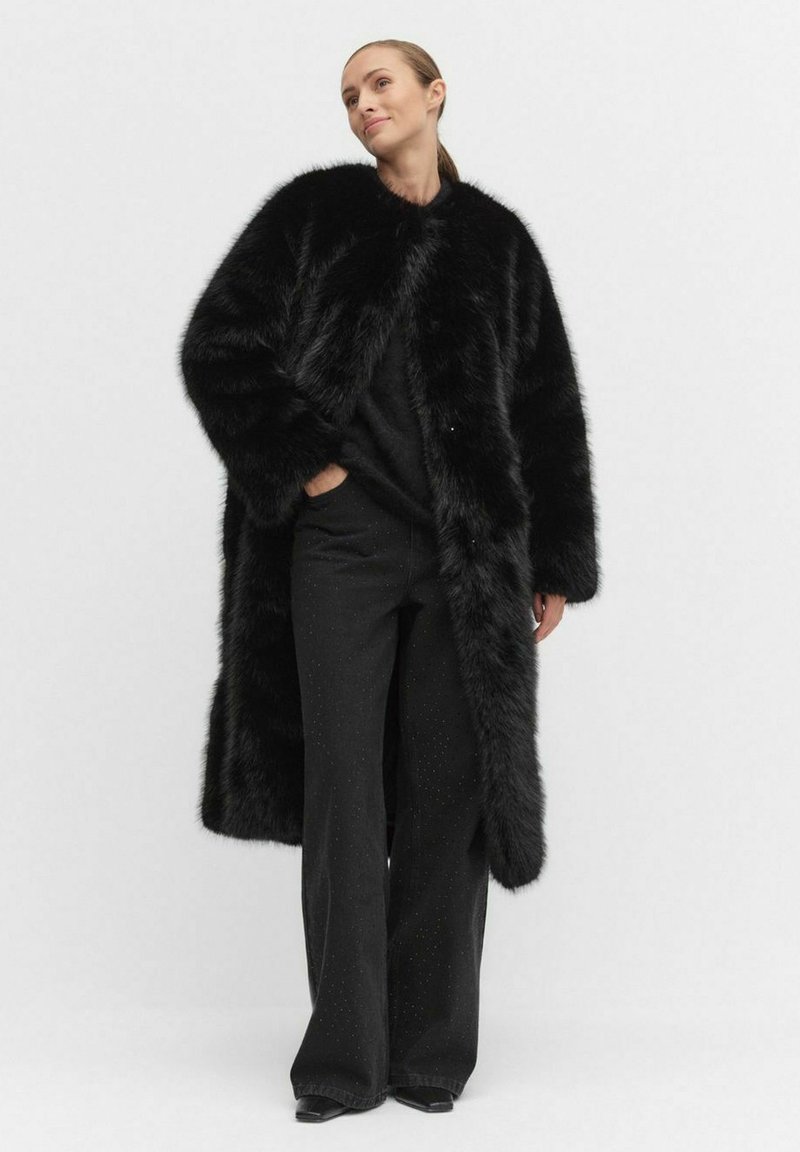 Musta keinoturkisviitta, jossa on pitkä ja valuva muotoilu. Siinä on pyöreä kaulus ja sivutaskut, yhdistettynä slim fit -mustiin housuihin.