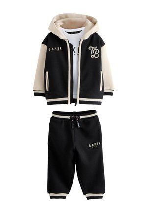 Młody chłopiec stoi, ubrany w czarną i beżową kurtkę z kapturem, białą koszulkę z napisem "BAKER", czarne spodnie oraz białe trampki.