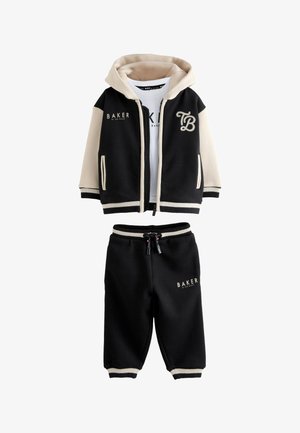 Młody chłopiec stoi, ubrany w czarną i beżową kurtkę z kapturem, białą koszulkę z napisem "BAKER", czarne spodnie oraz białe trampki.