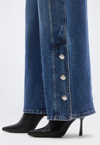 Μπλε τζιν παντελόνι denim με ασημένια κουμπιά σε όλο το εξωτερικό του πόδι, συνδυασμένο με μαύρα μπότακια με ψηλό τακούνι και μυτερή μύτη.