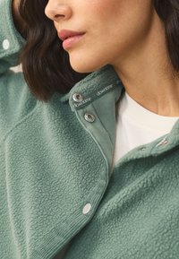 Giacca in pile verde menta con bottoni a pressione argentati e etichetta del colletto stampata. Presenta una superficie testurizzata e un design a strati.