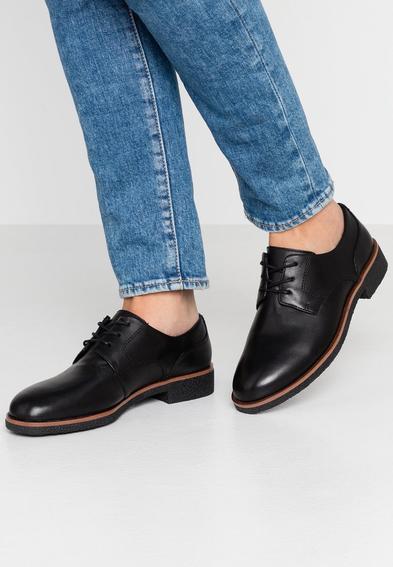 Chaussures en cuir noir à lacets portées avec un jean bleu, debout sur un sol gris clair avec un pied légèrement levé.