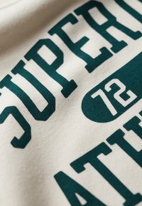 Kremowy materiał z wyraźnym zielonym napisem "SUPERIOR" i "72" w stylizowanej czcionce; miękka faktura z delikatnym połyskiem.