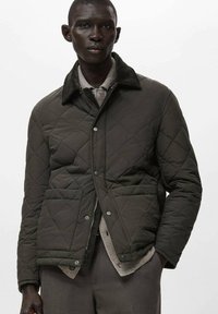 Giacca trapuntata verde scuro con colletto foderato in pelliccia nera, tasche anteriori e chiusura con zip, caratterizzata da un tessuto liscio e strutturato.