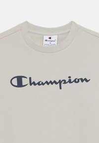 Lys beige bomull t-skjorte med en mørk marineblå 'Champion'-logo på brystet. Klassisk rund hals design, myk tekstur. Størrelse S-merkelapp synlig.