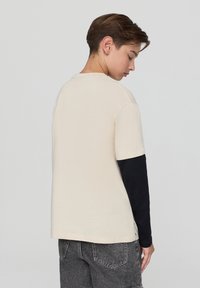Maglietta color crema a maniche corte indossata sopra un top a maniche lunghe nero. Texture morbida, taglio rilassato e dettagli sui fianchi.